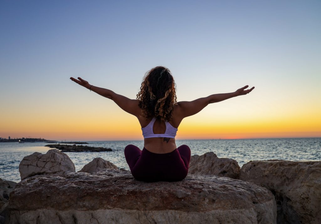 Woman sitting on rocks by the water at sunset, in a meditation position, legs crossed, reaching to the sky. 